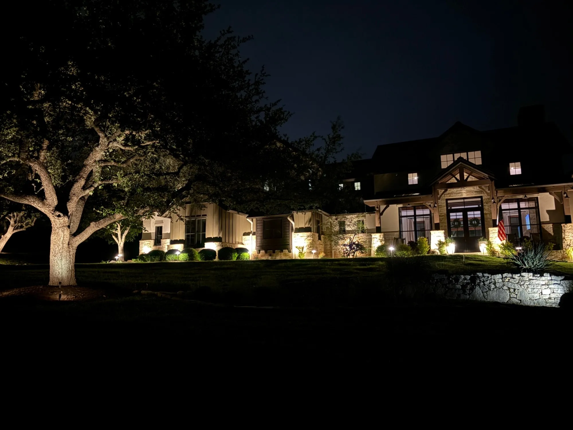 Hill Country home at night with uplit live oak, lit stone columns, path lights, and a glowing stone retaining wall