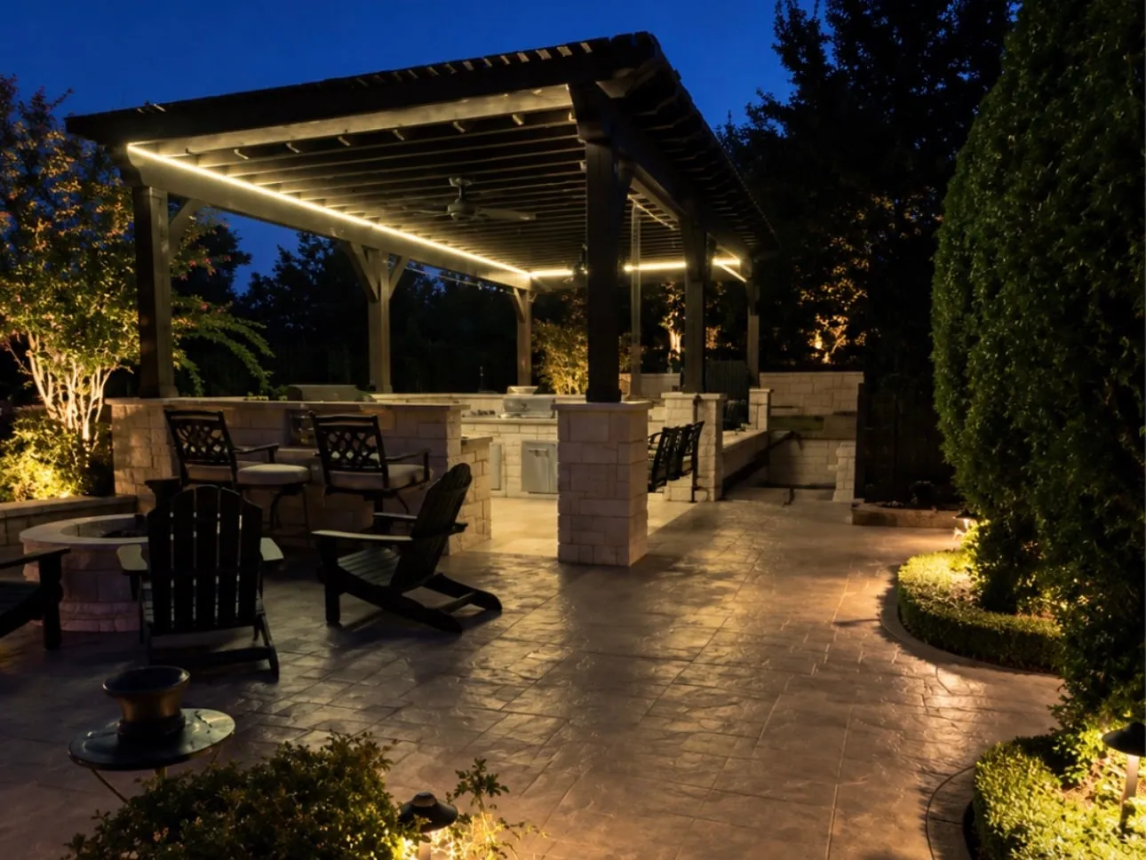 Pergola with LED strip lighting over an outdoor kitchen and fire pit, with path lights and uplit trees